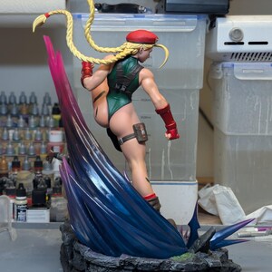 May include: A collectible figurine of a character with long blonde braided hair, wearing a green leotard, red beret, and red gauntlets. The figure is posed in a dynamic kick atop a dark, rocky base with a large, translucent blue and purple energy effect.