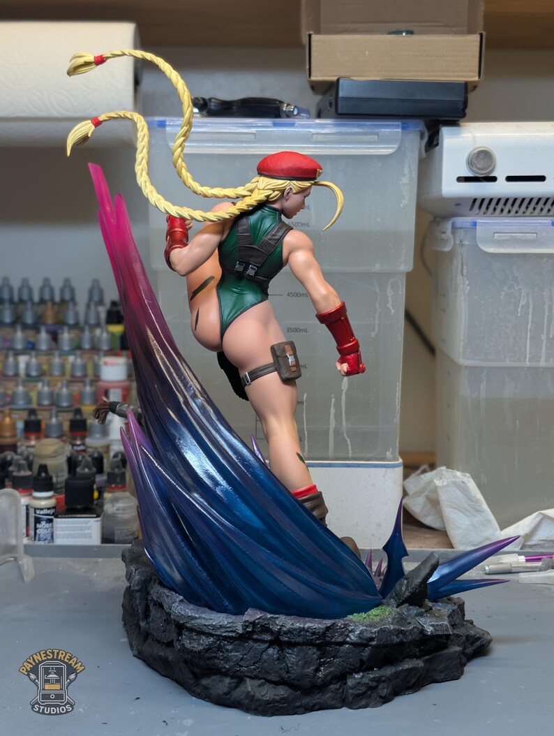 May include: A collectible figurine of a character with long blonde braided hair, wearing a green leotard, red beret, and red gauntlets. The figure is posed in a dynamic kick atop a dark, rocky base with a large, translucent blue and purple energy effect.