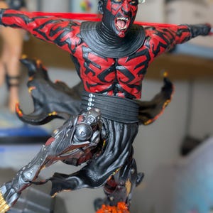 K&ouml;nnte beinhalten: Eine detaillierte Figur eines geh&ouml;rnten Charakters mit roten und schwarzen Hautmustern, offenem Mund und entbl&ouml;&szlig;ten Z&auml;hnen. Die Figur tr&auml;gt eine schwarze Tunika und eine metallische Beinr&uuml;stung und ist dynamisch &uuml;ber einem strukturierten orangefarbenen Sockel positioniert.