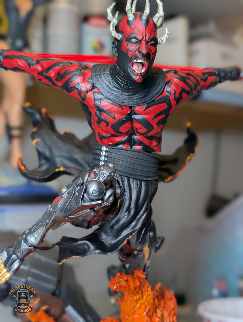 K&ouml;nnte beinhalten: Eine detaillierte Figur eines geh&ouml;rnten Charakters mit roten und schwarzen Hautmustern, offenem Mund und entbl&ouml;&szlig;ten Z&auml;hnen. Die Figur tr&auml;gt eine schwarze Tunika und eine metallische Beinr&uuml;stung und ist dynamisch &uuml;ber einem strukturierten orangefarbenen Sockel positioniert.