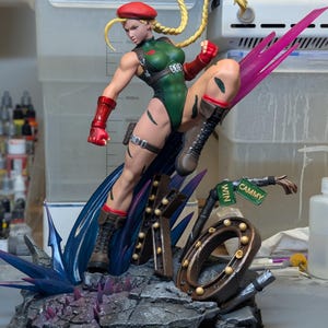 May include: A detailed figurine of the character Cammy from Street Fighter, wearing a green leotard, red beret, and red gauntlets. The figure is in a dynamic kicking pose atop a base featuring the letters KO, a street sign, and blue energy effects.