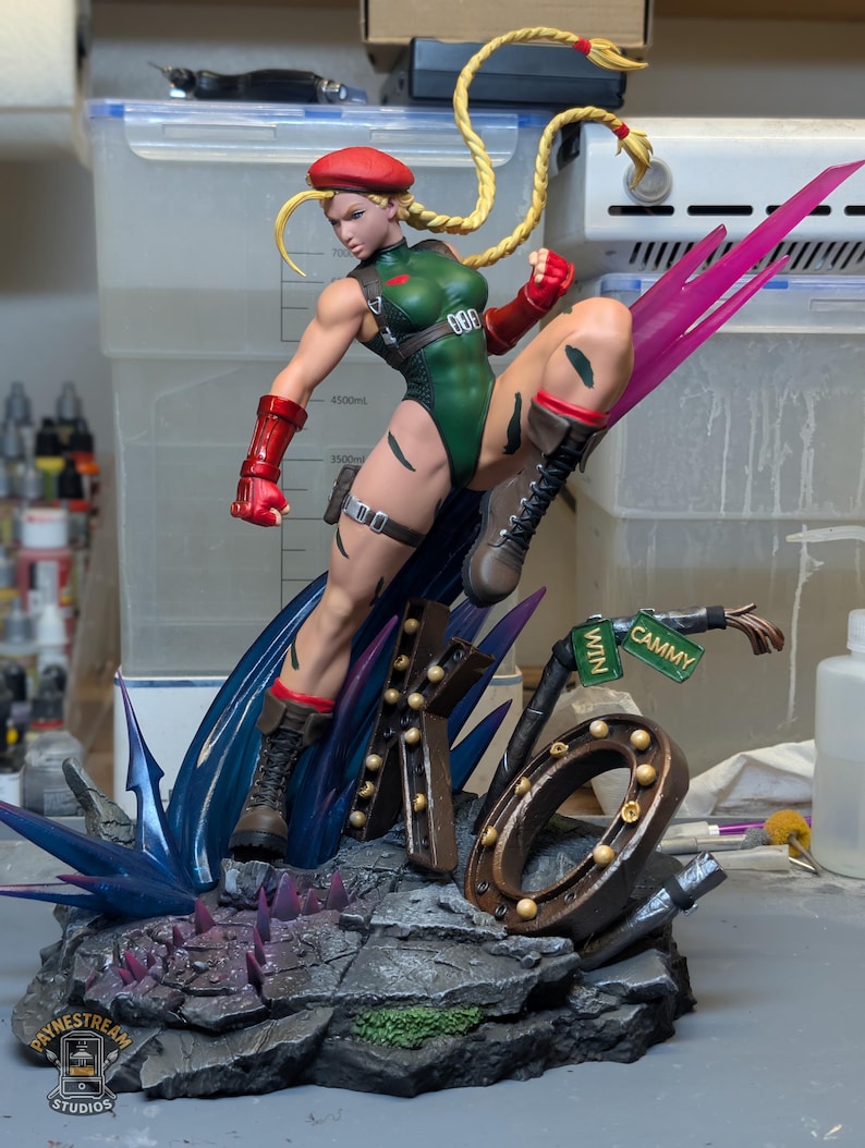 May include: A detailed figurine of the character Cammy from Street Fighter, wearing a green leotard, red beret, and red gauntlets. The figure is in a dynamic kicking pose atop a base featuring the letters KO, a street sign, and blue energy effects.