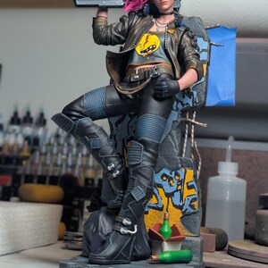 May include: A detailed collectible figurine of a character with bright pink hair, wearing a brown leather jacket, gray crop top with a yellow graphic, and black pants. The figure holds a black rifle and stands on a base with graffiti.
