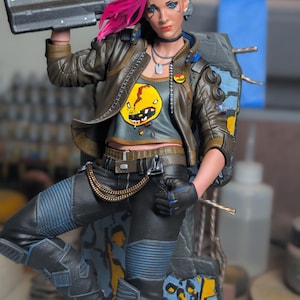 May include: A detailed action figure featuring a character with bright pink hair, wearing a brown leather jacket over a gray crop top with a yellow graphic print. The figure holds a black futuristic rifle and wears black pants with blue accents and a gold chain.