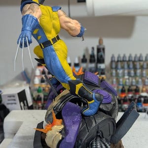 May include: A detailed figurine of Wolverine in a dynamic pose, wearing his classic yellow and blue costume. The figure has sharp claws extended and is positioned atop a detailed, multi-colored base. The figurine is a collectible item.