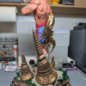 May include: A muscular figurine standing on a base with two bronze-colored bell-shaped structures. The figure wears blue shorts with red side stripes and has a gold-colored flowing element attached to the side. The base features green accents.