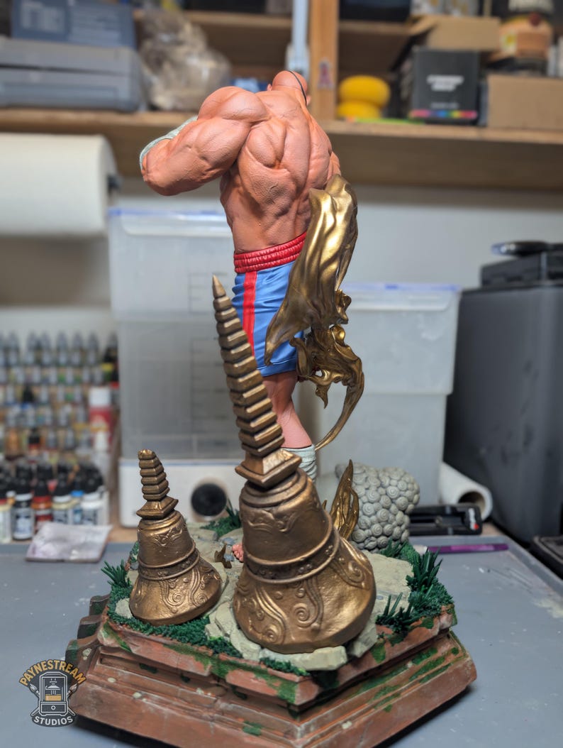 May include: A muscular figurine standing on a base with two bronze-colored bell-shaped structures. The figure wears blue shorts with red side stripes and has a gold-colored flowing element attached to the side. The base features green accents.
