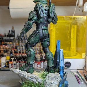 May include: A green and black armored soldier figurine standing on a rocky base with green grass details. The figure wears a helmet with a gold visor and carries a long weapon on its back. A blue metallic structure is attached to the base.
