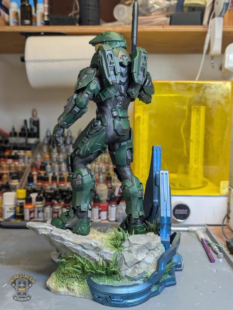 May include: A green and black armored soldier figurine standing on a rocky base with green grass details. The figure wears a helmet with a gold visor and carries a long weapon on its back. A blue metallic structure is attached to the base.