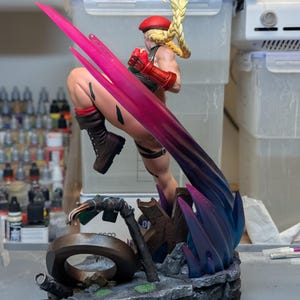 May include: A painted figurine of a blonde character with a long braided ponytail wearing a red beret, green leotard, and brown boots. The character is in a dynamic kicking pose with a translucent pink energy effect and stands on a rocky base.