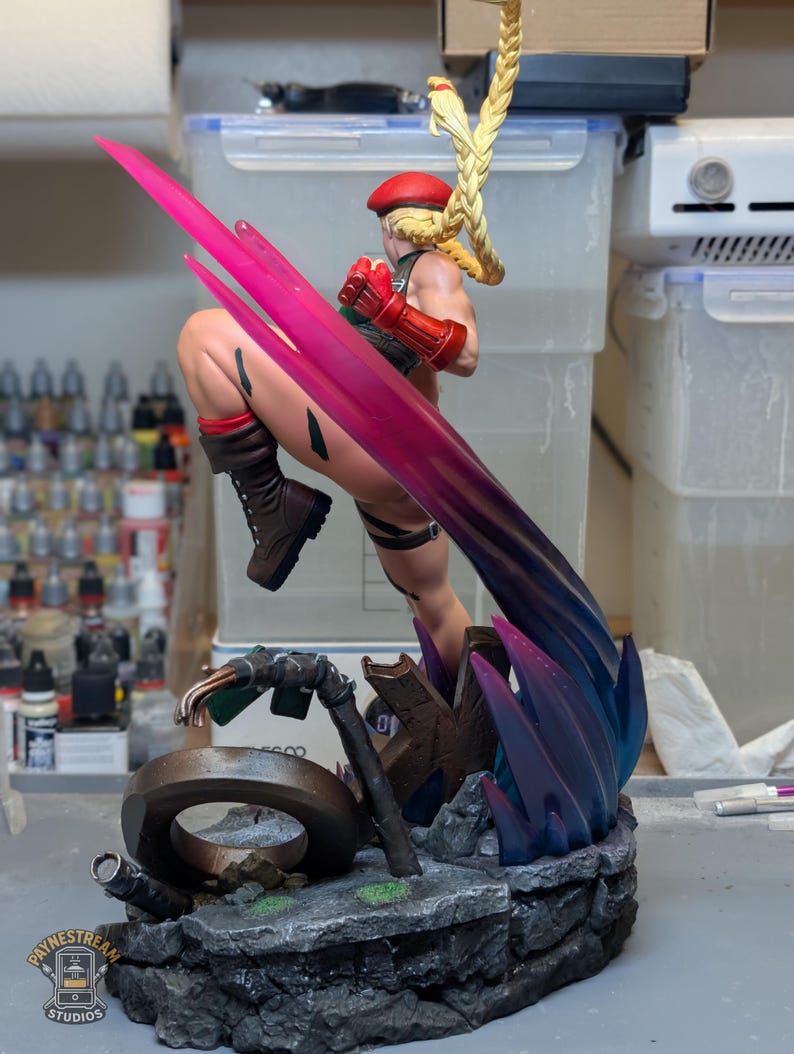 May include: A painted figurine of a blonde character with a long braided ponytail wearing a red beret, green leotard, and brown boots. The character is in a dynamic kicking pose with a translucent pink energy effect and stands on a rocky base.