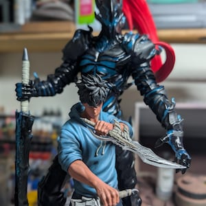 May include: A detailed figurine set featuring a dark armored warrior with a tall red ponytail standing behind a figure in a light blue hoodie. The warrior holds a long black sword, while the figure in the hoodie wields two silver-toned blades.