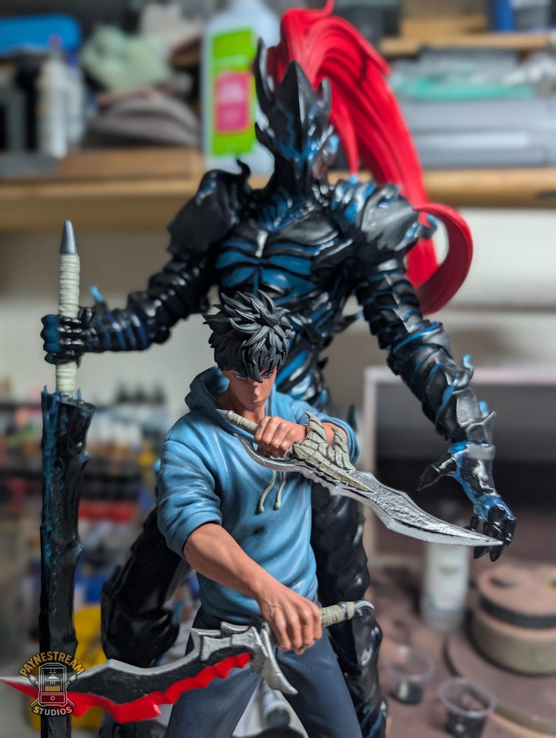 May include: A detailed figurine set featuring a dark armored warrior with a tall red ponytail standing behind a figure in a light blue hoodie. The warrior holds a long black sword, while the figure in the hoodie wields two silver-toned blades.