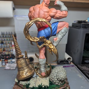 May include: A muscular, bald action figure in a dynamic fighting pose, wearing blue shorts and white hand and leg wraps. The figure is surrounded by a swirling gold energy effect, standing on a detailed base with a gold bell and stone textures.