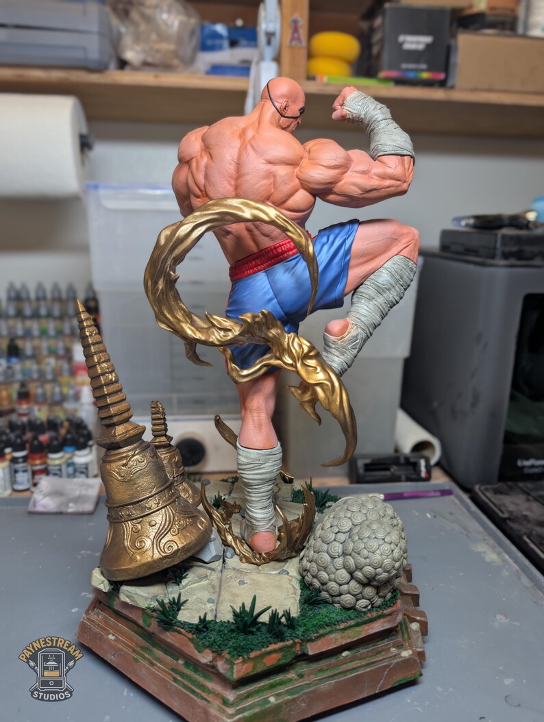 May include: A muscular, bald action figure in a dynamic fighting pose, wearing blue shorts and white hand and leg wraps. The figure is surrounded by a swirling gold energy effect, standing on a detailed base with a gold bell and stone textures.