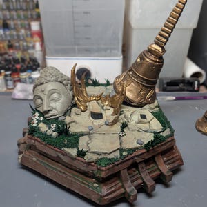May include: A diorama base featuring a stone Buddha head, a tall golden spire, and golden flame accents set upon a cracked stone floor with green moss and small white flowers. The base has a brown wooden-style frame with a Paynestream Studios logo.
