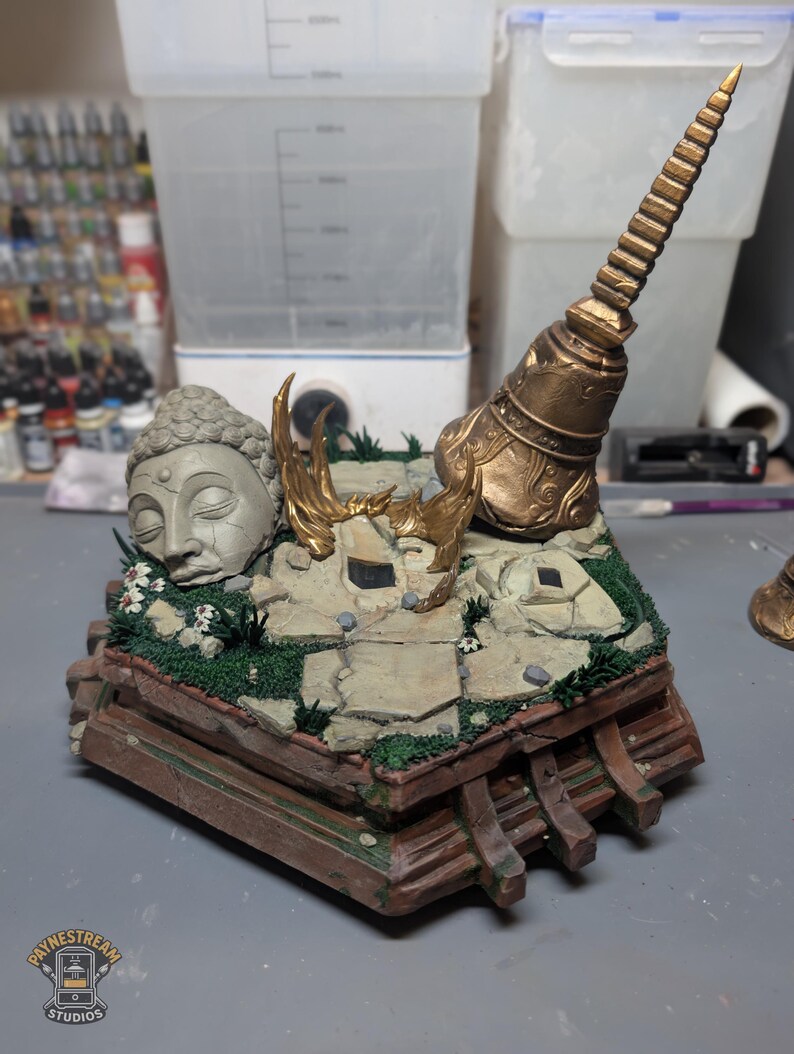 May include: A diorama base featuring a stone Buddha head, a tall golden spire, and golden flame accents set upon a cracked stone floor with green moss and small white flowers. The base has a brown wooden-style frame with a Paynestream Studios logo.