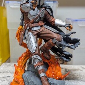 K&ouml;nnte beinhalten: Detaillierte Figur von The Mandalorian in dynamischer Pose, mit einem Blaster. Die Figur hat einen silbernen Helm, braune und silberne R&uuml;stung und steht vor einem Hintergrund aus stilisierten orangefarbenen Flammen und einem felsigen Sockel. Die Figur ist etwa 30 cm gro&szlig;.
