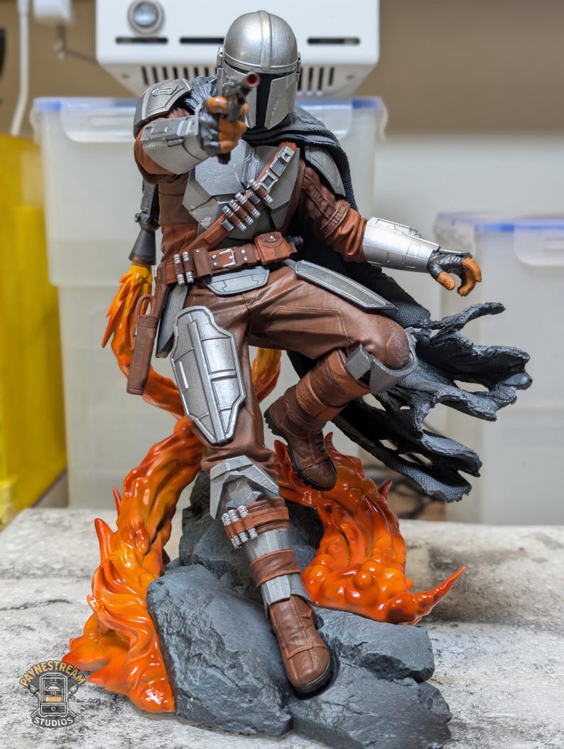 K&ouml;nnte beinhalten: Detaillierte Figur von The Mandalorian in dynamischer Pose, mit einem Blaster. Die Figur hat einen silbernen Helm, braune und silberne R&uuml;stung und steht vor einem Hintergrund aus stilisierten orangefarbenen Flammen und einem felsigen Sockel. Die Figur ist etwa 30 cm gro&szlig;.
