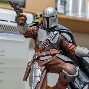 K&ouml;nnte beinhalten: Detaillierte Figur von The Mandalorian in dynamischer Pose, die einen Blaster h&auml;lt. Die Figur hat einen silbernen Helm, braune und graue R&uuml;stung und einen Umhang. Eine kleine, gr&uuml;ne Grogu-Figur sitzt auf der Schulter. Die Figur steht auf einem Sockel.