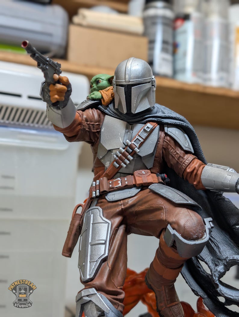 K&ouml;nnte beinhalten: Detaillierte Figur von The Mandalorian in dynamischer Pose, die einen Blaster h&auml;lt. Die Figur hat einen silbernen Helm, braune und graue R&uuml;stung und einen Umhang. Eine kleine, gr&uuml;ne Grogu-Figur sitzt auf der Schulter. Die Figur steht auf einem Sockel.