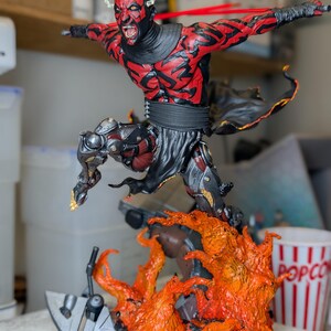 K&ouml;nnte beinhalten: Eine detaillierte Actionfigur eines rot-schwarzen geh&ouml;rnten Charakters mit grimmigem Gesichtsausdruck, der &uuml;ber einen modellierten Sockel aus orangefarbenen und gelben Flammen springt. Die Figur tr&auml;gt schwarze Gew&auml;nder und eine metallische Beinar&uuml;stung.