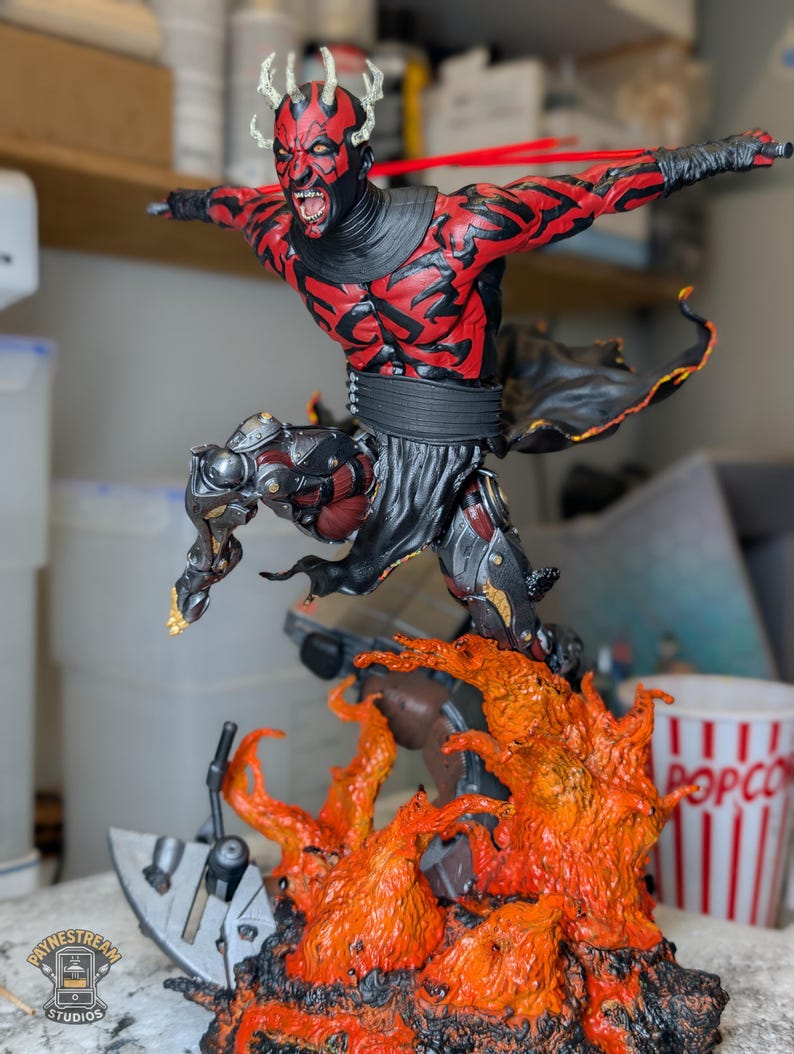 K&ouml;nnte beinhalten: Eine detaillierte Actionfigur eines rot-schwarzen geh&ouml;rnten Charakters mit grimmigem Gesichtsausdruck, der &uuml;ber einen modellierten Sockel aus orangefarbenen und gelben Flammen springt. Die Figur tr&auml;gt schwarze Gew&auml;nder und eine metallische Beinar&uuml;stung.
