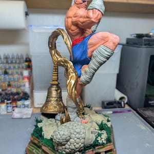 May include: A highly detailed, hand-painted resin statue of a muscular fighter in a dynamic kicking pose. The figure wears blue shorts and white hand wraps, standing on a base featuring a golden bell, a stone head sculpture, and green grass.