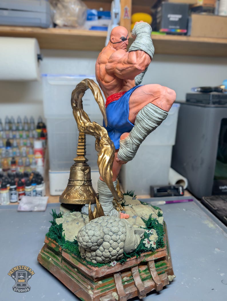 May include: A highly detailed, hand-painted resin statue of a muscular fighter in a dynamic kicking pose. The figure wears blue shorts and white hand wraps, standing on a base featuring a golden bell, a stone head sculpture, and green grass.