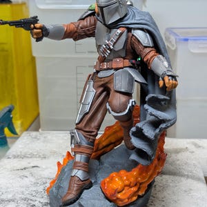 K&ouml;nnte beinhalten: Detaillierte Actionfigur von The Mandalorian, in silberner und brauner R&uuml;stung, mit einem Blaster. Die Figur steht auf einem felsigen Sockel mit orangefarbenen Lava-Akzenten. Ein grauer Umhang bedeckt die Schultern.