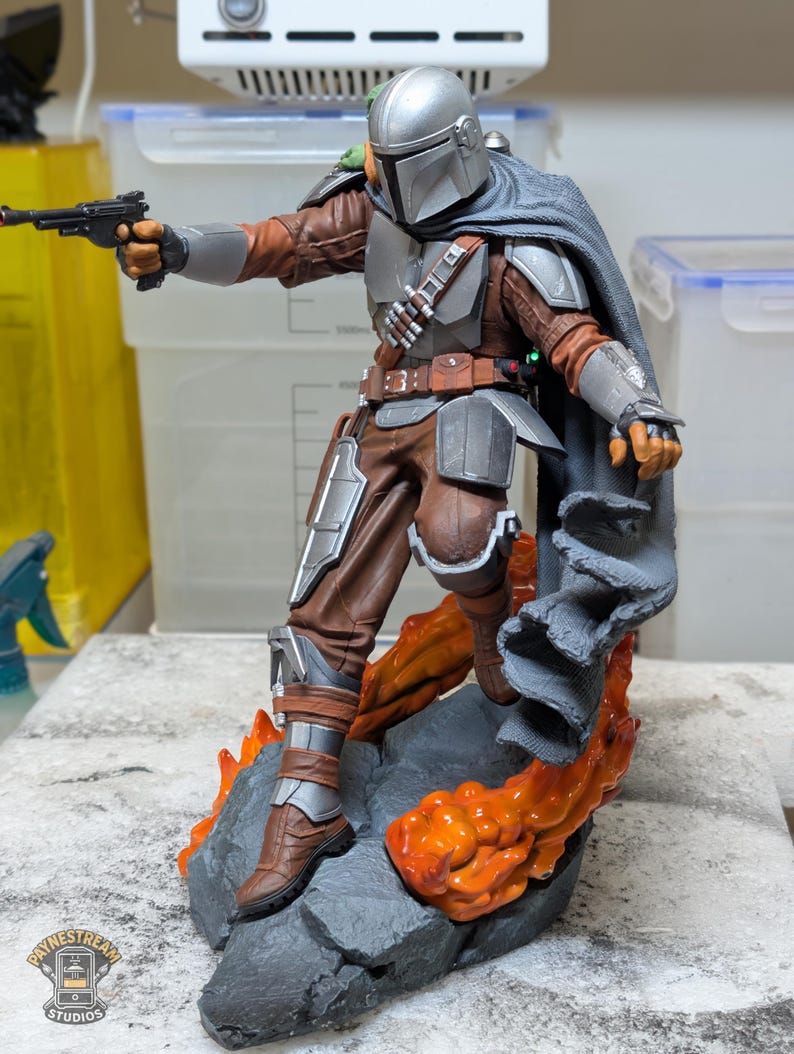 K&ouml;nnte beinhalten: Detaillierte Actionfigur von The Mandalorian, in silberner und brauner R&uuml;stung, mit einem Blaster. Die Figur steht auf einem felsigen Sockel mit orangefarbenen Lava-Akzenten. Ein grauer Umhang bedeckt die Schultern.