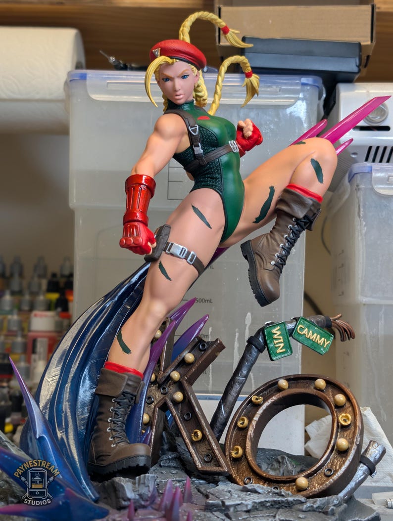 May include: A detailed figurine of a blonde character in a green leotard, red beret, and brown combat boots, posed in a dynamic kick. The base features a large metallic KO sign with small lights and two green signs labeled WIN and CAMMY.