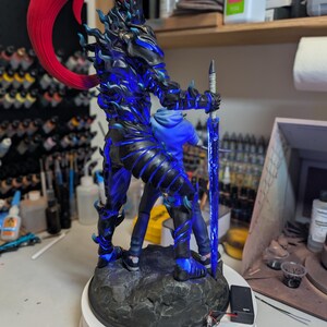 May include: A detailed figurine featuring a dark, armored character with a vibrant red ponytail and glowing blue accents, holding a long, translucent blue sword. The figure stands on a textured, rocky base with an attached battery pack and wires.