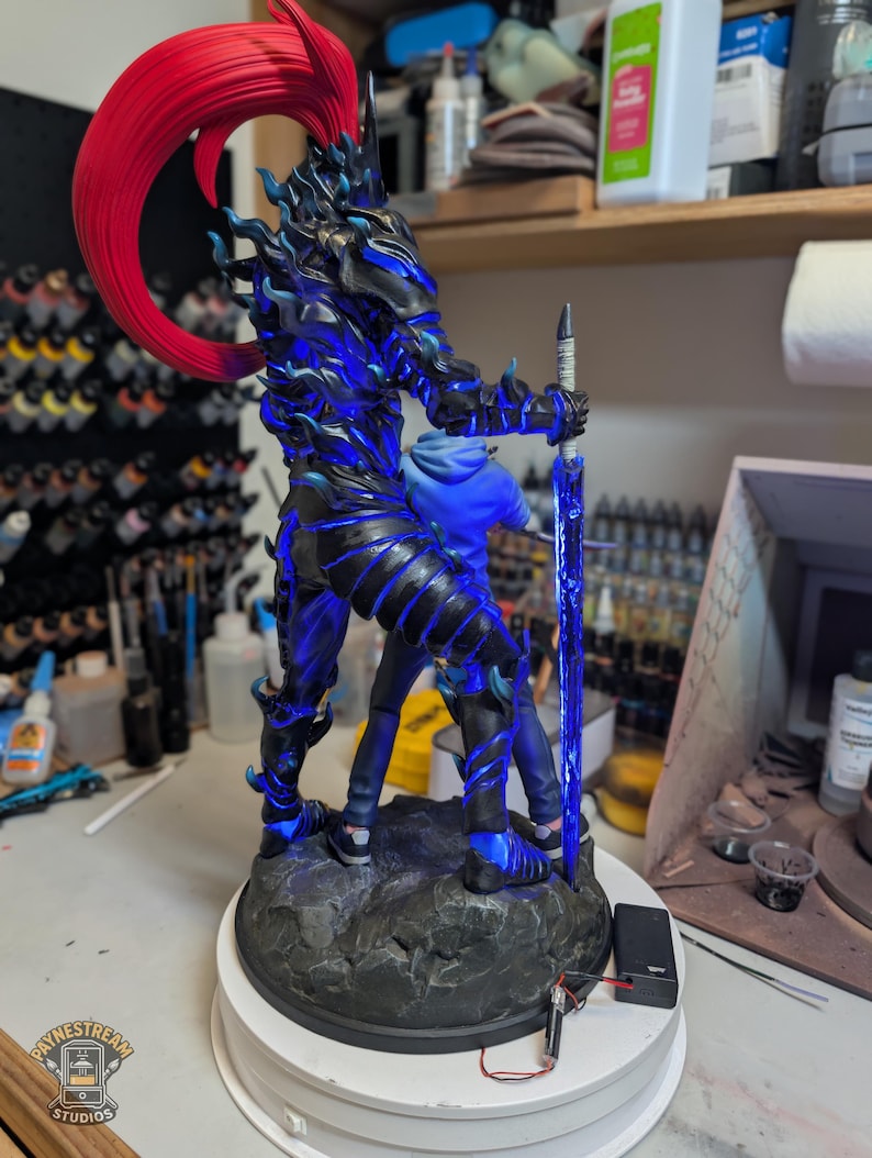 May include: A detailed figurine featuring a dark, armored character with a vibrant red ponytail and glowing blue accents, holding a long, translucent blue sword. The figure stands on a textured, rocky base with an attached battery pack and wires.