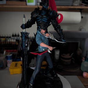 May include: A detailed figurine featuring a character in a blue hoodie and dark pants holding two daggers, standing in front of a tall, dark, armored shadow figure with a red ponytail and a large black sword with blue glowing accents.