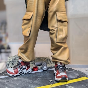 May include: A figurine featuring tan cargo pants with side pockets and red, white, and black sneakers with the text "KILLER BEE" on the side. The figure stands on a circular base designed to look like a cracked asphalt street with a yellow line.