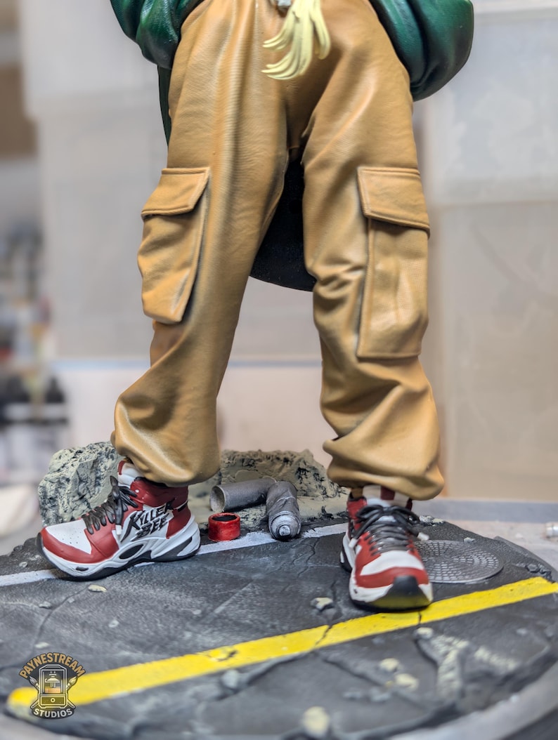 May include: A figurine featuring tan cargo pants with side pockets and red, white, and black sneakers with the text "KILLER BEE" on the side. The figure stands on a circular base designed to look like a cracked asphalt street with a yellow line.