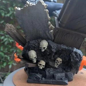 May include: A detailed statue featuring a dark, textured base with several small skulls. The statue has a dark color scheme with hints of orange and gold. The base has a wooden appearance, and the statue is set on a round, light brown surface.