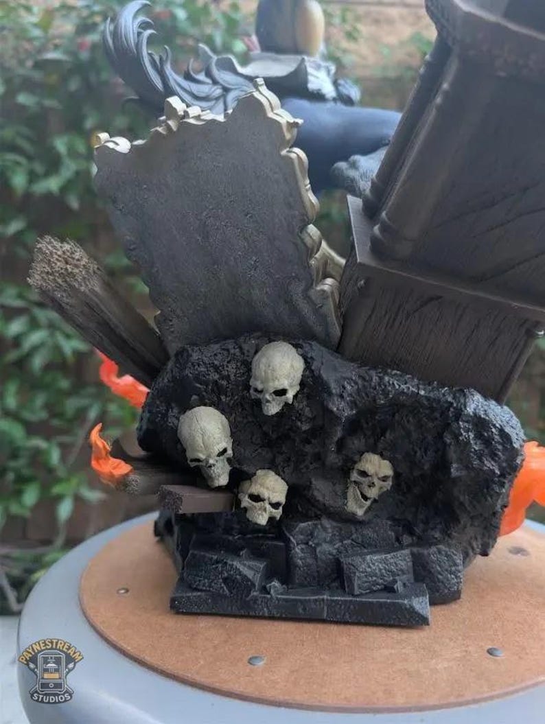 May include: A detailed statue featuring a dark, textured base with several small skulls. The statue has a dark color scheme with hints of orange and gold. The base has a wooden appearance, and the statue is set on a round, light brown surface.