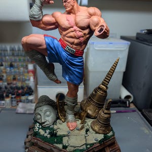 May include: A muscular male figurine with a scarred chest and eye patch, wearing blue boxing shorts with the word TIGER on the waistband. The figure is posed in a fighting stance on a base featuring a stone Buddha head and golden bells.