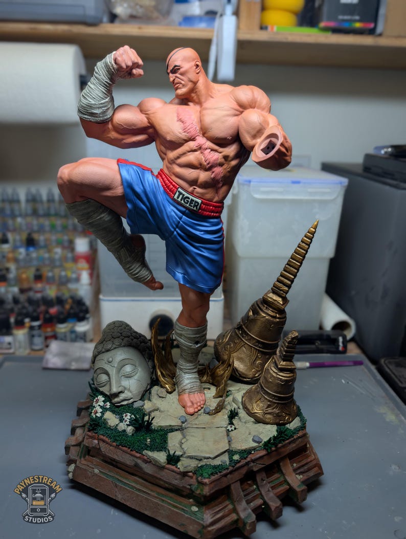 May include: A muscular male figurine with a scarred chest and eye patch, wearing blue boxing shorts with the word TIGER on the waistband. The figure is posed in a fighting stance on a base featuring a stone Buddha head and golden bells.