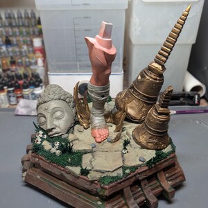 May include: A painted diorama base featuring a stone-colored Buddha head, a single human leg wrapped in bandages, and two ornate gold-colored temple structures. The scene is set on a cracked stone floor with patches of green grass and small white flowers.