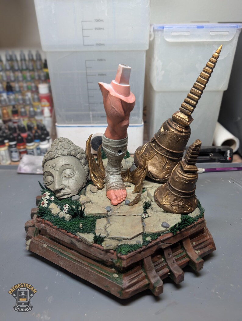 May include: A painted diorama base featuring a stone-colored Buddha head, a single human leg wrapped in bandages, and two ornate gold-colored temple structures. The scene is set on a cracked stone floor with patches of green grass and small white flowers.