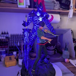 May include: A detailed figurine featuring a young person in a green hoodie and dark pants standing in front of a large, dark, skeletal shadow figure with glowing blue accents and a red ponytail, holding a large sword and two smaller daggers.
