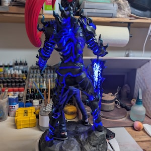 May include: A standing figurine of a humanoid character with a large red ponytail, dark armor featuring glowing blue energy patterns, and a large sword held in the right hand. The figure is mounted on a textured gray rock-like base.