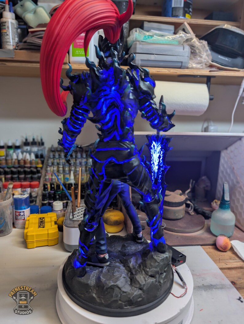 May include: A standing figurine of a humanoid character with a large red ponytail, dark armor featuring glowing blue energy patterns, and a large sword held in the right hand. The figure is mounted on a textured gray rock-like base.