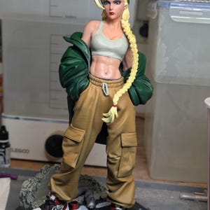May include: A collectible figurine of a blonde character with a long braid wearing a green beanie, a light gray sports bra, a dark green jacket, and tan cargo pants. The character wears red and white sneakers and stands on a circular base.
