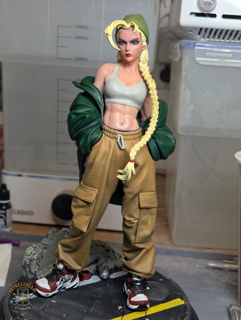 May include: A collectible figurine of a blonde character with a long braid wearing a green beanie, a light gray sports bra, a dark green jacket, and tan cargo pants. The character wears red and white sneakers and stands on a circular base.