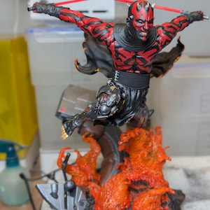 K&ouml;nnte beinhalten: Eine detaillierte Actionfigur eines rot-schwarz t&auml;towierten Charakters mit H&ouml;rnern, der &uuml;ber einer Basis aus geformten orangefarbenen und schwarzen Flammen springt. Die Figur h&auml;lt ein rotes Doppellichtschwert horizontal und tr&auml;gt eine schwarze Tunika.