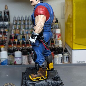 May include: A detailed figurine of a character in a red cap, red shirt, and blue overalls, with a mustache. The figure is standing on a gray base, with a black gun and white gloves. The figure is a stylized representation of a video game character.